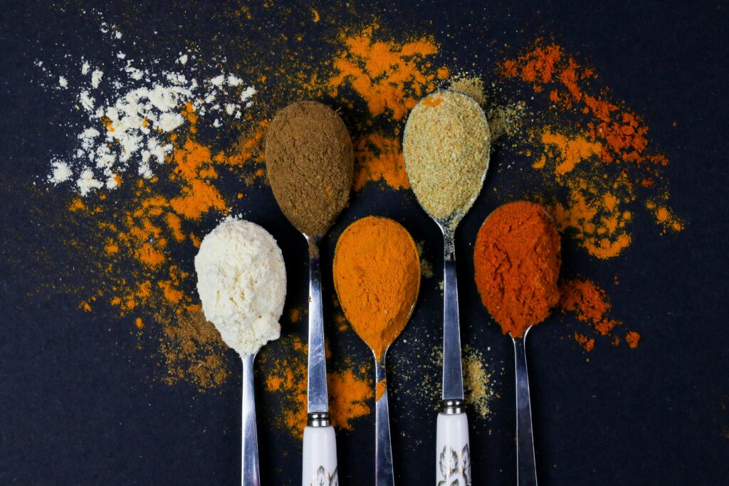 Colorful spices displayed on spoons with scattered powders, showcasing culinary variety.